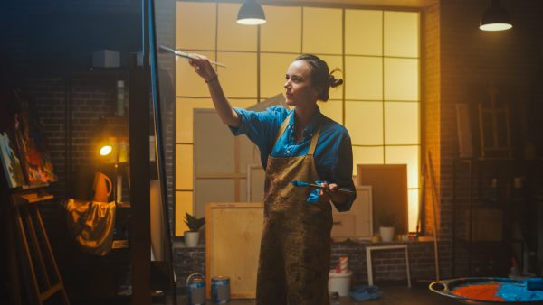 Portrait of Talented Female Artist Working on a Modern Abstract Oil Painting, Gesturing with Broad Strokes Using Paint Brush. Dark Creative Studio Large Picture Stands on Easel Illuminated. Low Angle