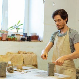 bright-studio.-concentrated-bearded-guy-being-a-professional-pottery-master-and-separating-piece-of-clay-with-wire