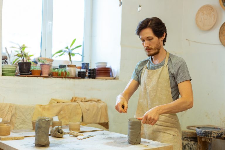 bright-studio.-concentrated-bearded-guy-being-a-professional-pottery-master-and-separating-piece-of-clay-with-wire