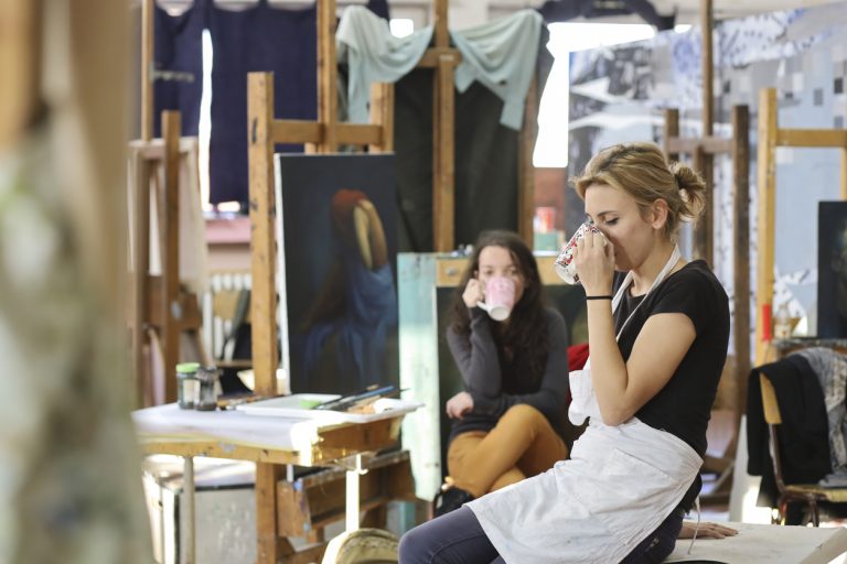 woman-artistis-sitting-and-working-on-the-paitings-and-drinking-coffee-in-a-messy-atelier