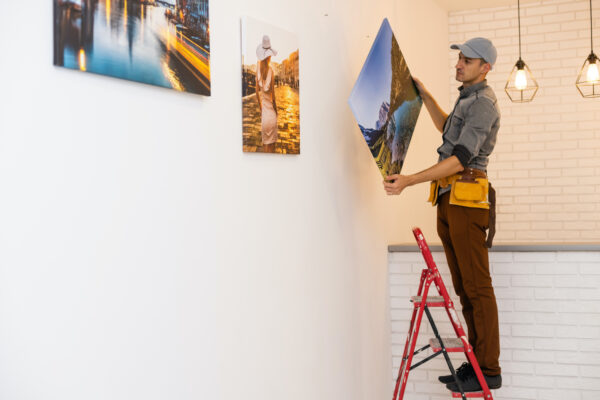 Man on a ladder hanging photography artwork on a white office wall