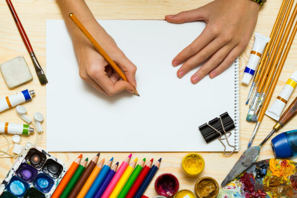 Image of hands of a women artist about to draw on a blank page with colorful art supplies surrounding the paper.