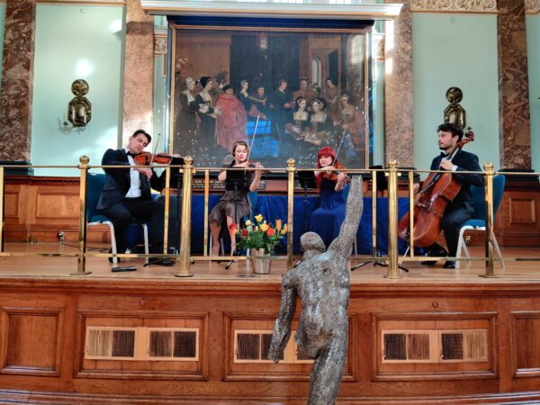 4-person orchestra playing in front of a painting and behind a sculpture at the London Art Biennale