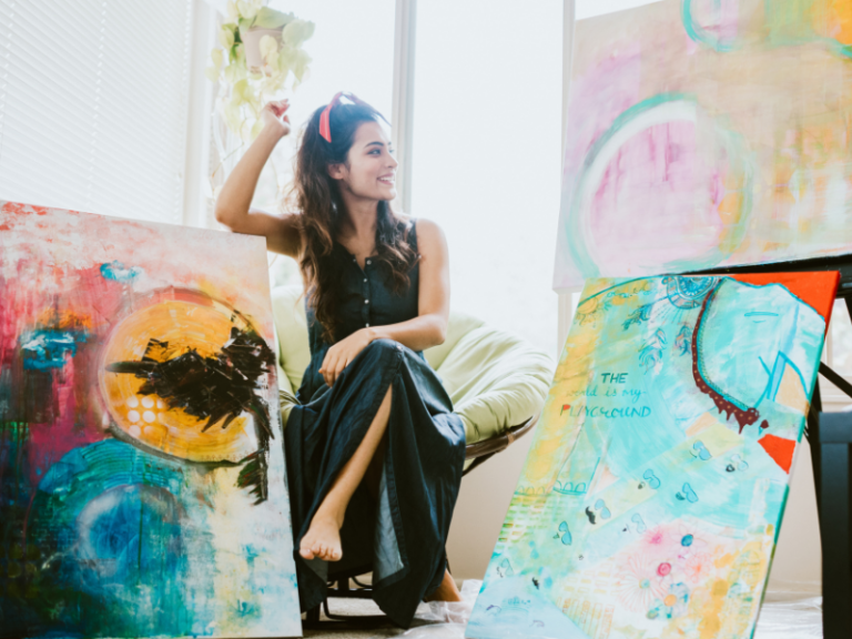 artist sitting in studio surrounded by three colorful artworks.