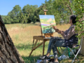 women, seated in a chair in a field near a tree, en plein air painting on an easel