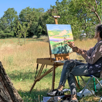 women, seated in a chair in a field near a tree, en plein air painting on an easel